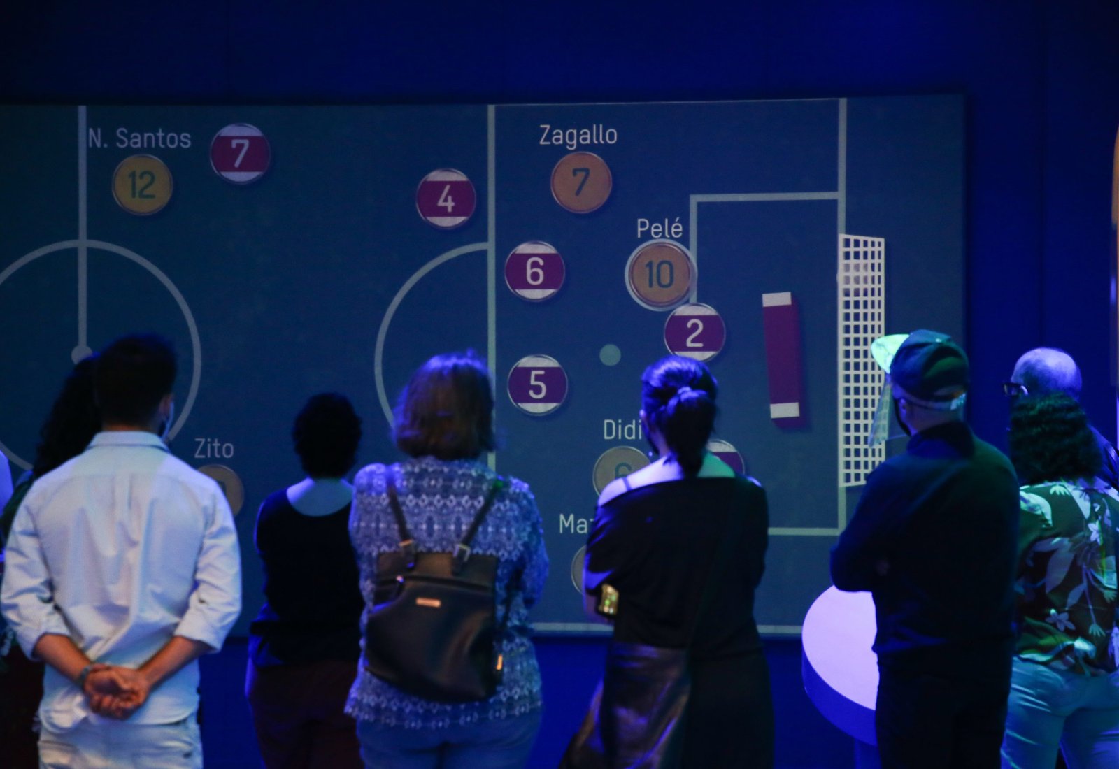 Pessoas de costas assistem numa tela um vídeo com um jogo de botão.