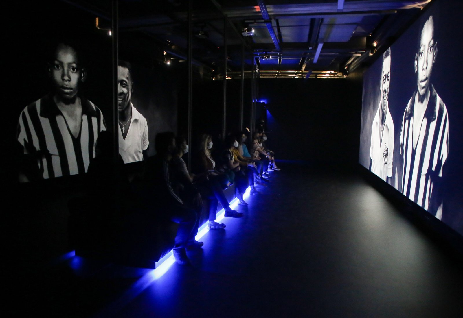 Numa sala escura, pessoas assistem um filme numa telão. O banco onde estão sentadas tem uma luz azul por baixo. Na tela, vemos uma foto de Pelé adolescente.