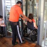 Funcionário do Educativo do Museu auxiliando uma mulher com cadeira de rodas a entrar no elevador enquanto outro rapaz também de cadeira de rodas, segura a porta do elevador