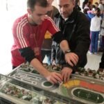 Três pessoas na foto. Um educador auxiliando um outro rapaz a tocar na maquete do estádio do Pacaembu com crianças no fundo.
