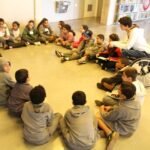 CriCrianças sentadas em círculo ouvindo atentamente uma mulher falando.