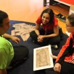 Três pessoas sentadas no chão, uma delas mostra uma planta do estádio do Pacaembu enquanto um homem e uma mulher ouvem atentamente.