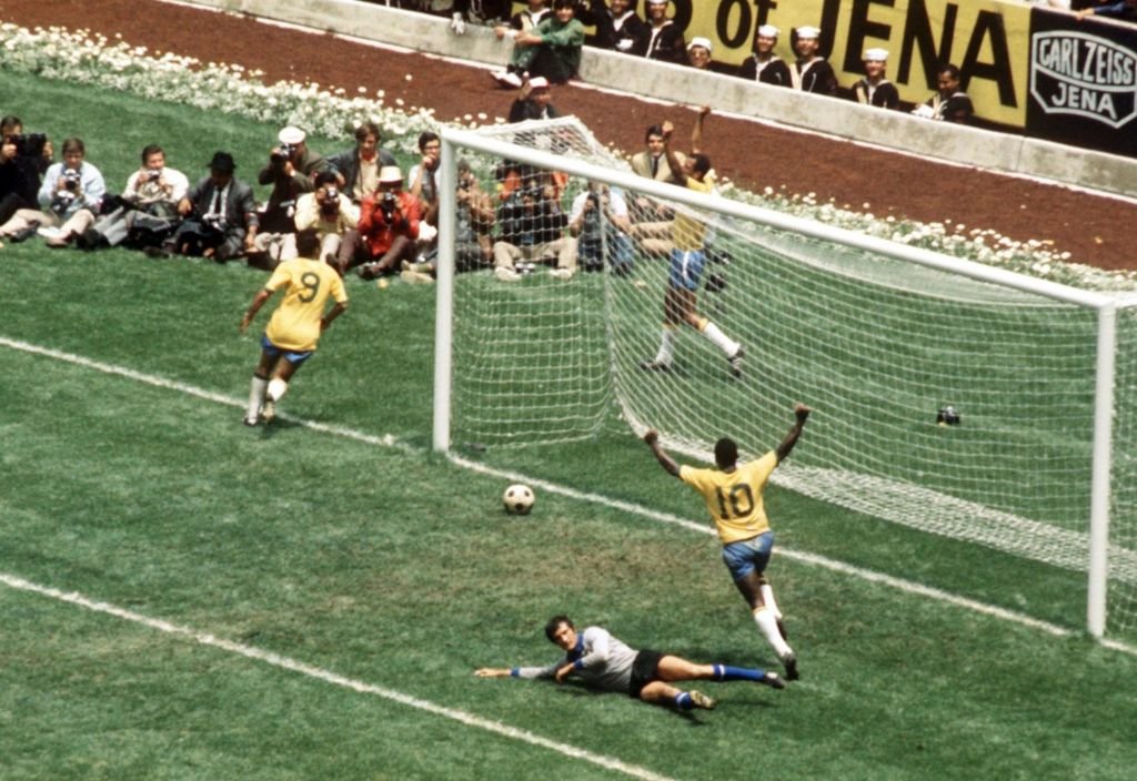 Pelé de costas levanta os braços em comemoração a um gol, com a bola ainda por passar a linha. O goleiro adversário está no chão.