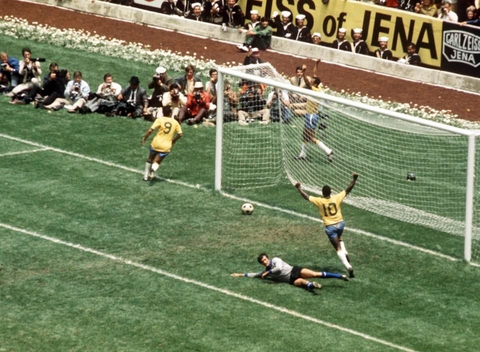 Pelé de costas levanta os braços em comemoração a um gol, com a bola ainda por passar a linha. O goleiro adversário está no chão.