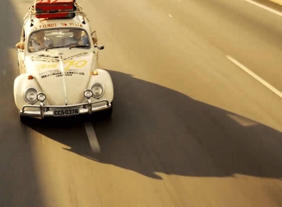 Um fusca branco fotografado na estrada, com sua sombra projetada no asfalto, num fim de tarde.