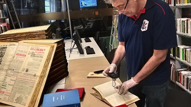 Um homem de pé fotografa a página de um livro. Ele usa luvas.