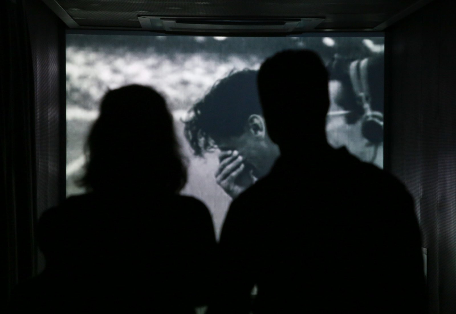 IMG_6664 Sala Rito de Passagem: um homem e uma mulher de costas, em contraluz, observam de pé a imagem projetada na parede à frente deles de um homem chorando com a mão no rosto.