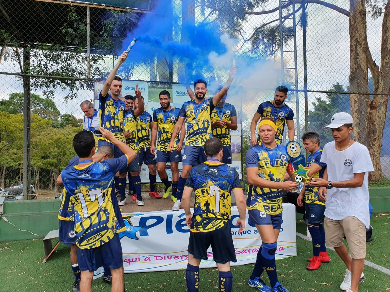 MF0000009308i Um grupo de rapazes com uniforme azul e amarelo comemora um título em um pódio. Eles estão alegres e alguns têm nas mãos sinalizadores que soltam fumaça azul.