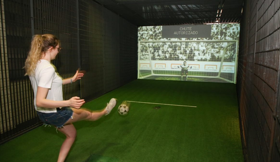 Uma moça chuta a bola em direção a um gol, defendido por um goleiro eletrônico