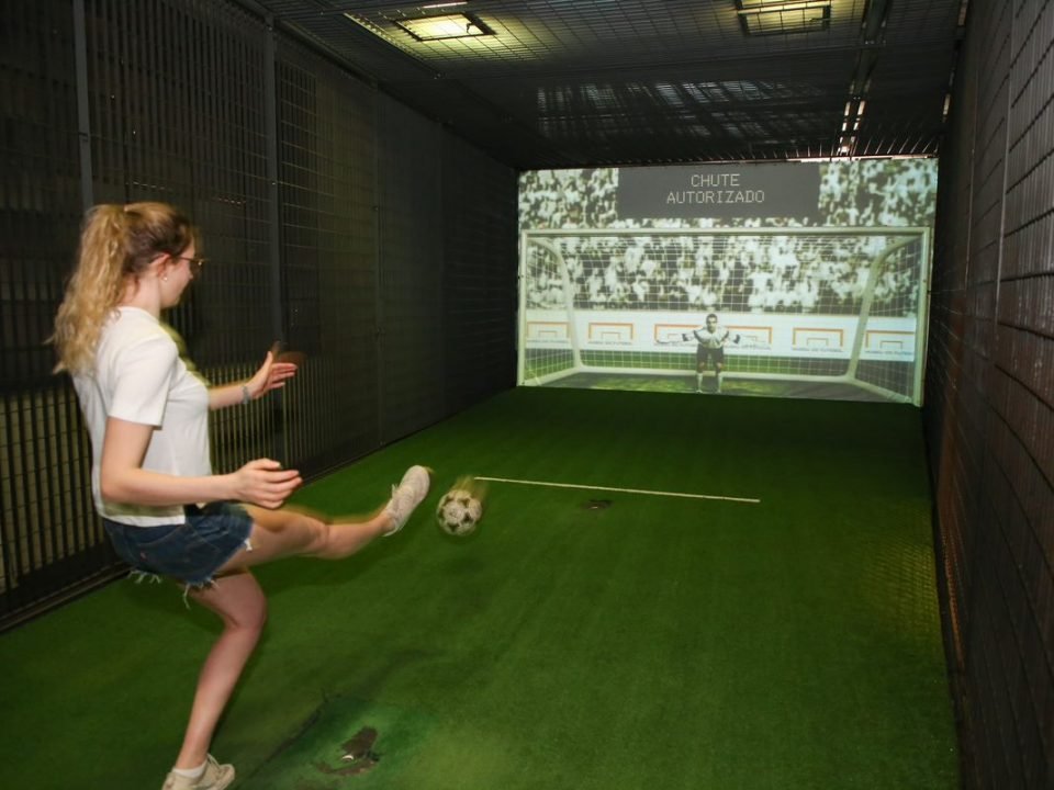 Uma moça chuta a bola em direção a um gol, defendido por um goleiro eletrônico