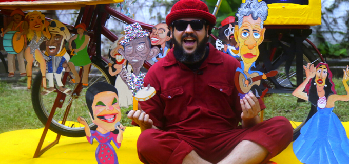 Um homem de barba e vestido inteiramente na cor vinho, inlcuindo um chapéu coco, está sentando no chão com vários personagens cortados em papel nas mãos e ao seu lado. Atrás dele há uma bicicleta.