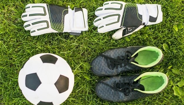 Sobre um gramado, há uma bola, um par de chuteiras pretas e um par de luvas brancas de goleiro.