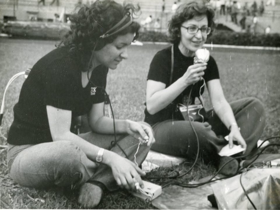 Foto em preto e branco de duas mulheres sentadas em um gramado, sendo que uma delas fala ao microfone de um gravador.