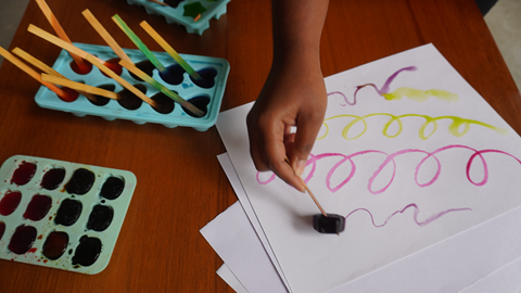 Uma mão aparece desenhando em papel com um dos gelos coloridos, segurando pelo palito.