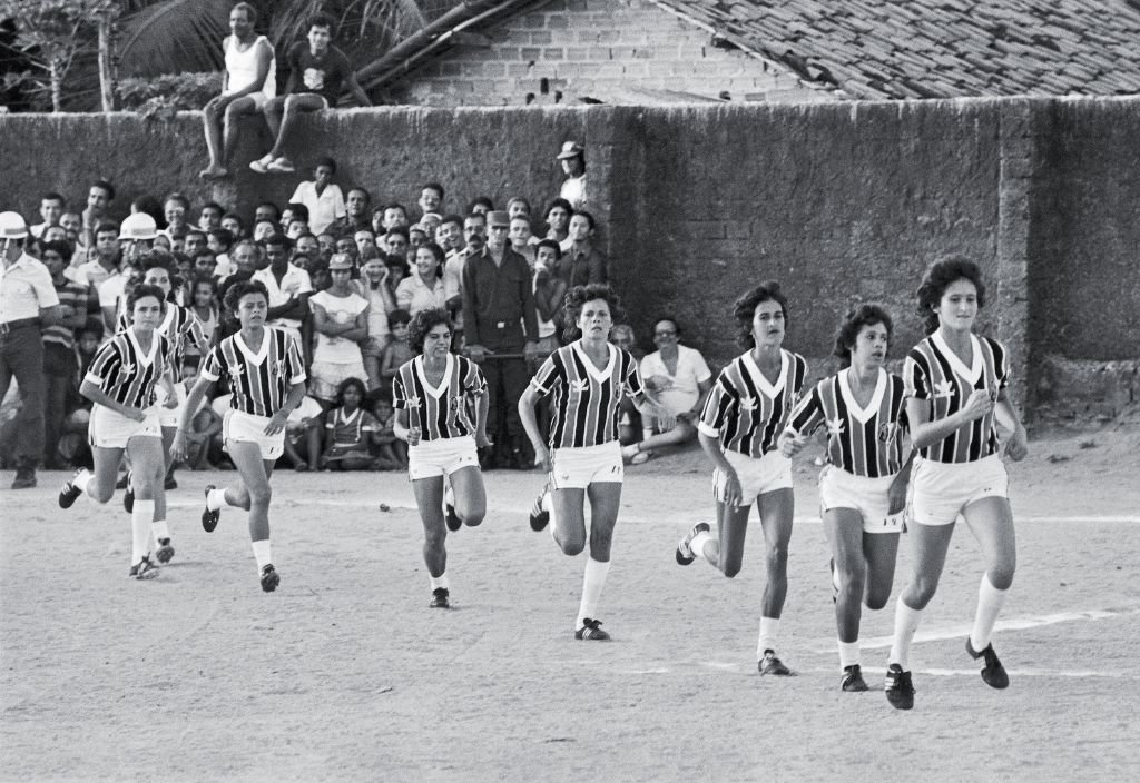 Imagem em preto e branco de mulheres vestidas com uniforme de futebol e torcedores ao fundo