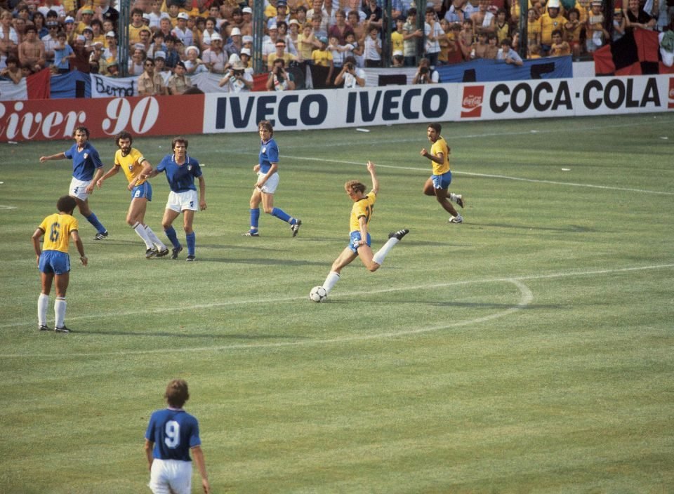 Foto de um estádio, torcedores ao fundo com camisa amarelo e branco, assistindo uma partida de futebol.