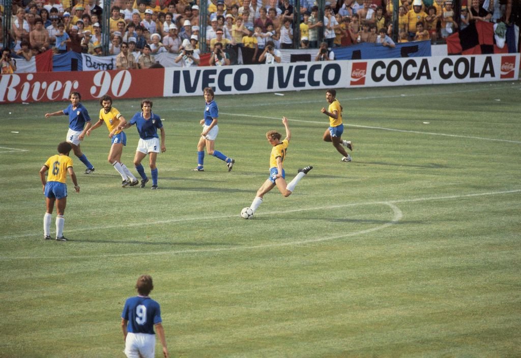 Foto de um estádio, torcedores ao fundo com camisa amarelo e branco, assistindo uma partida de futebol.