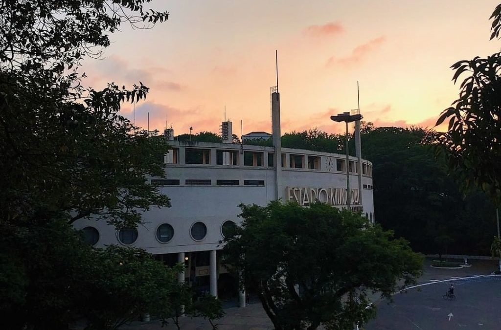 Imagem da fachada do Estádio do Pacaembu com um céu alaranjado e árvores