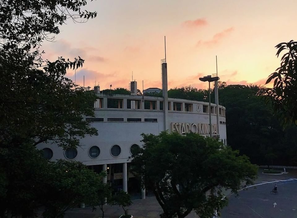 Imagem da fachada do Estádio do Pacaembu com um céu alaranjado e árvores