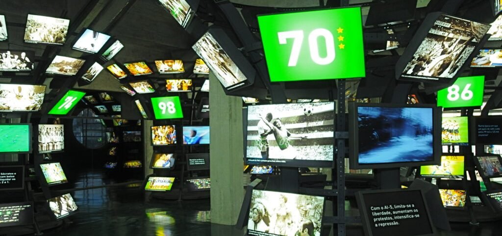 A foto mostra a Sala das Copas do mundo, com várias telas coloridas e preto e branco com fotos