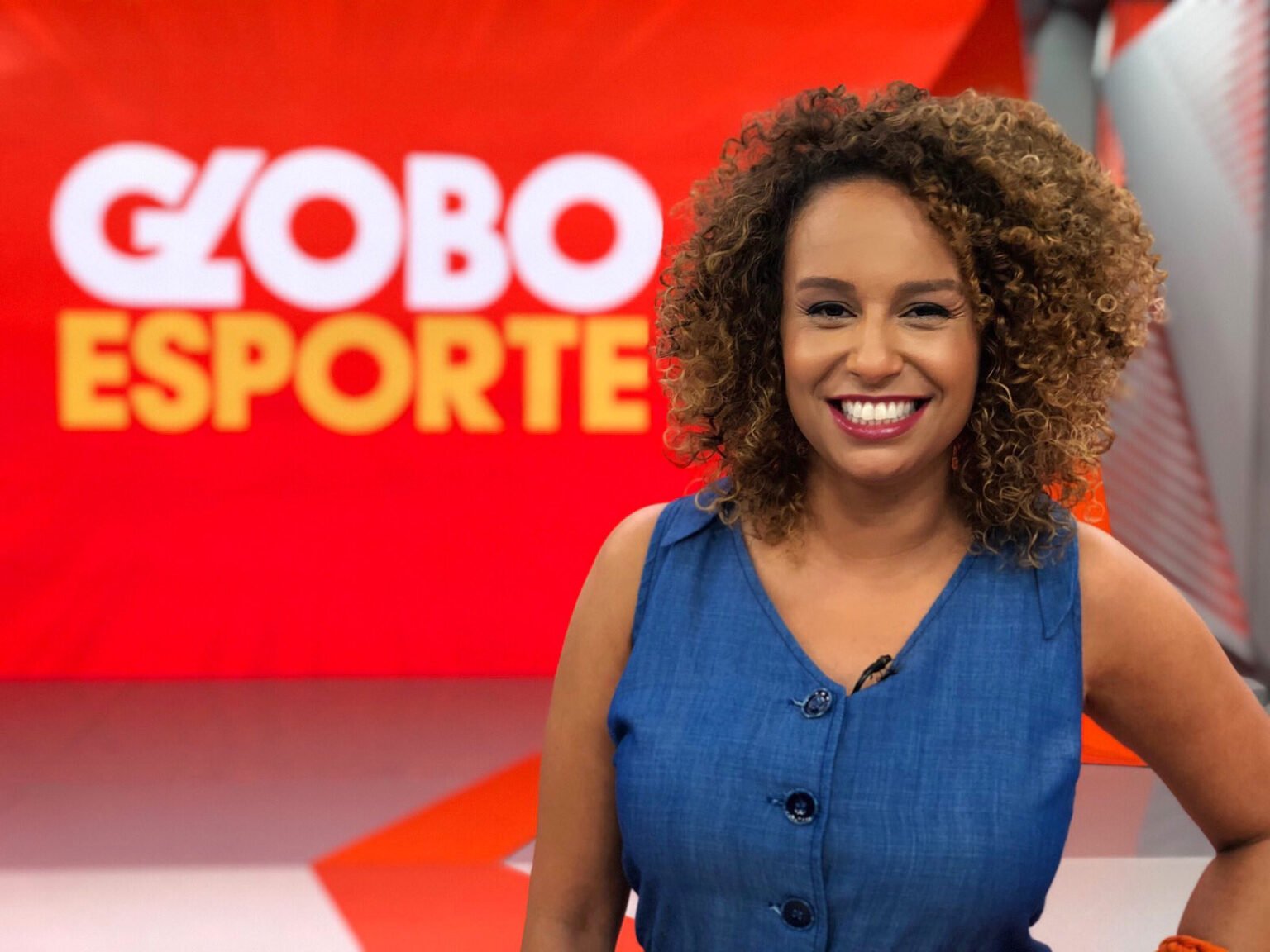 Karine Alves. Site Dibradoras. Karine é uma mulher negra, de cabelos cacheados. Ela uma uma blusa azul escura e sorri para a câmera. No fundo, o cenário vermelho do Globo Esporte