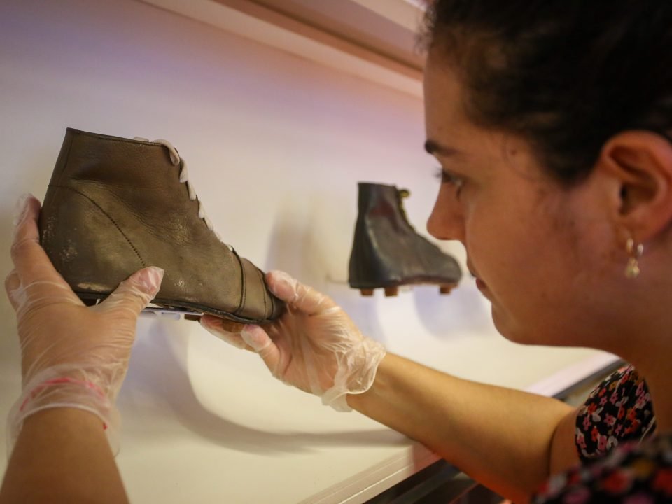 Foto de uma museóloga retirando uma chuteira antiga de couro de uma vitrine. Ela usa luvas.
