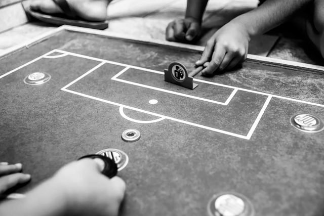 FOTO_CarolSantos - Caroline Lopes Imagem em preto e branco de uma mesa de botão. Vemos as mãos de uma pessoa atacando e de outra manejando um goleiro de plástico.