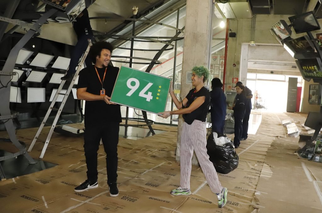 Um funcionário e uma funcionária do Museu do Futebol carregam uma placa verde com 94 escrito e quatro estrelas douradas,
