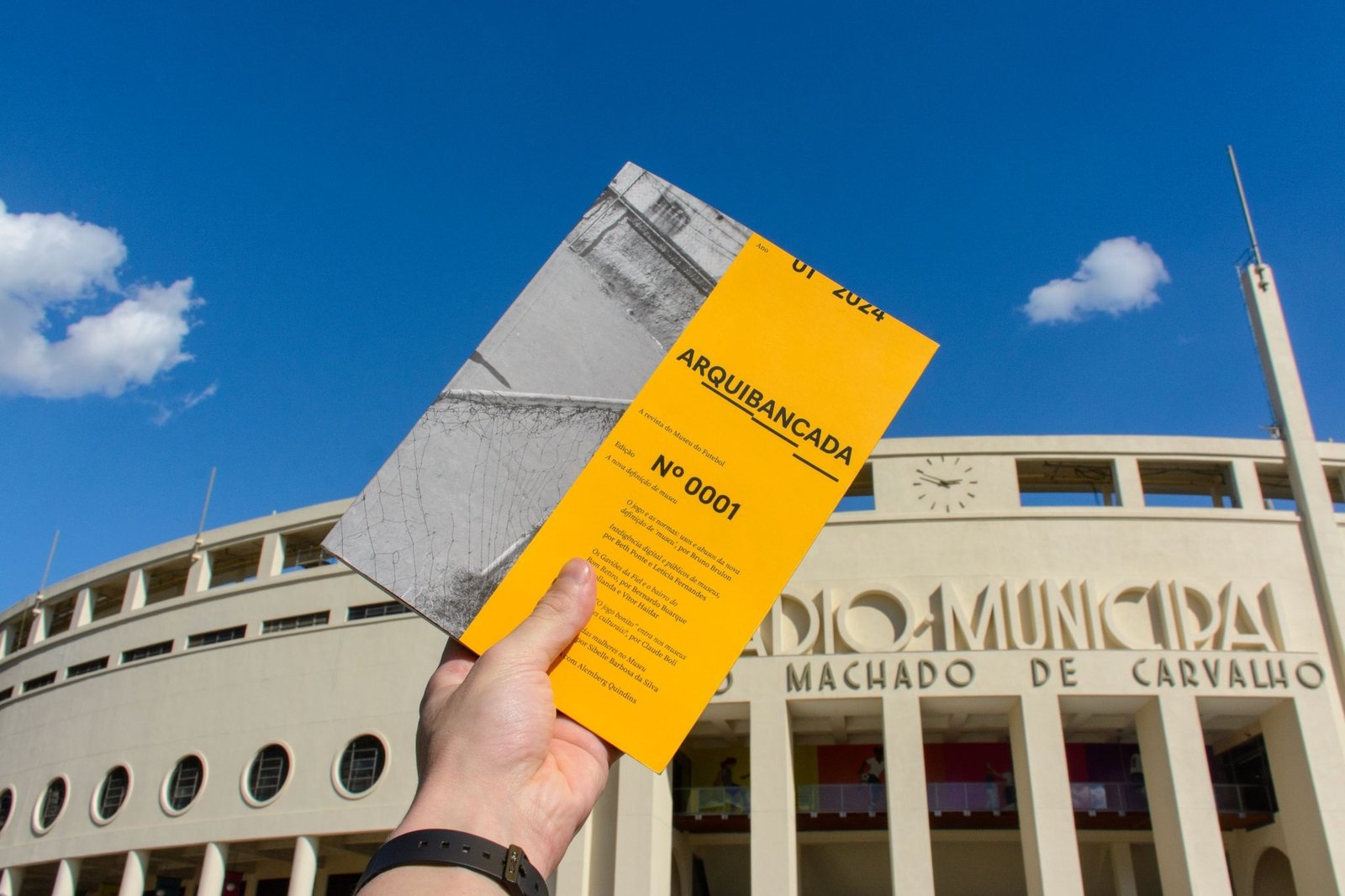 Imagem com o Estádio do Pacaembu ao fundo e uma mão segurando a Revista Arquibancada