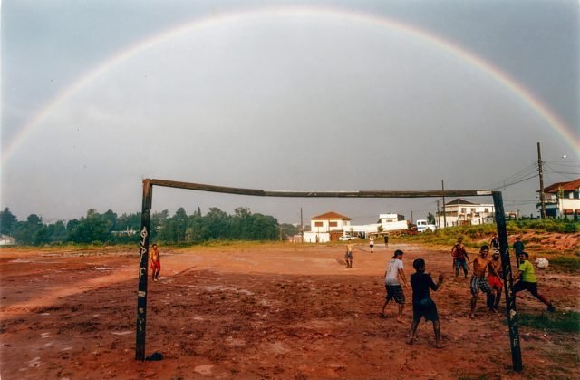 PauloPinto Um arco-íris compõe o cenário de crianças jogando futebol em um campo cheio de lama. A trave é improvisada e as crianças observam o destino da bola.