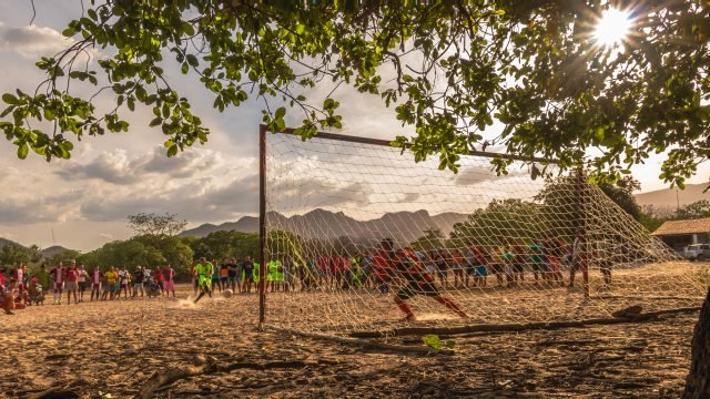 PedroJorgeDoNascimento Em um campo lotado torcedores e jogadores assistem ao atleta batendo um pênalti. O campo é de areia e iluminado pelo sol.