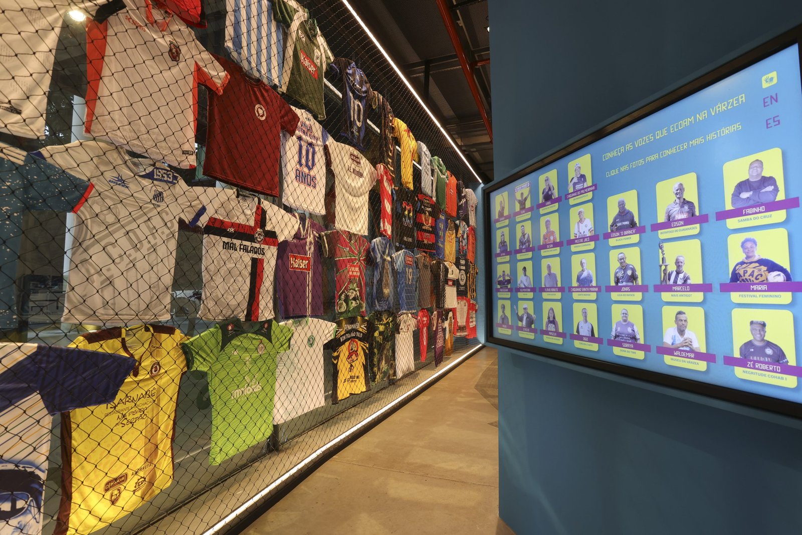 MF_Expo_Vozes_20241128_027 Tela de TV com as fotos e nomes de várias pessoas da várzea. No fundo, um varal com camisas de várzea.