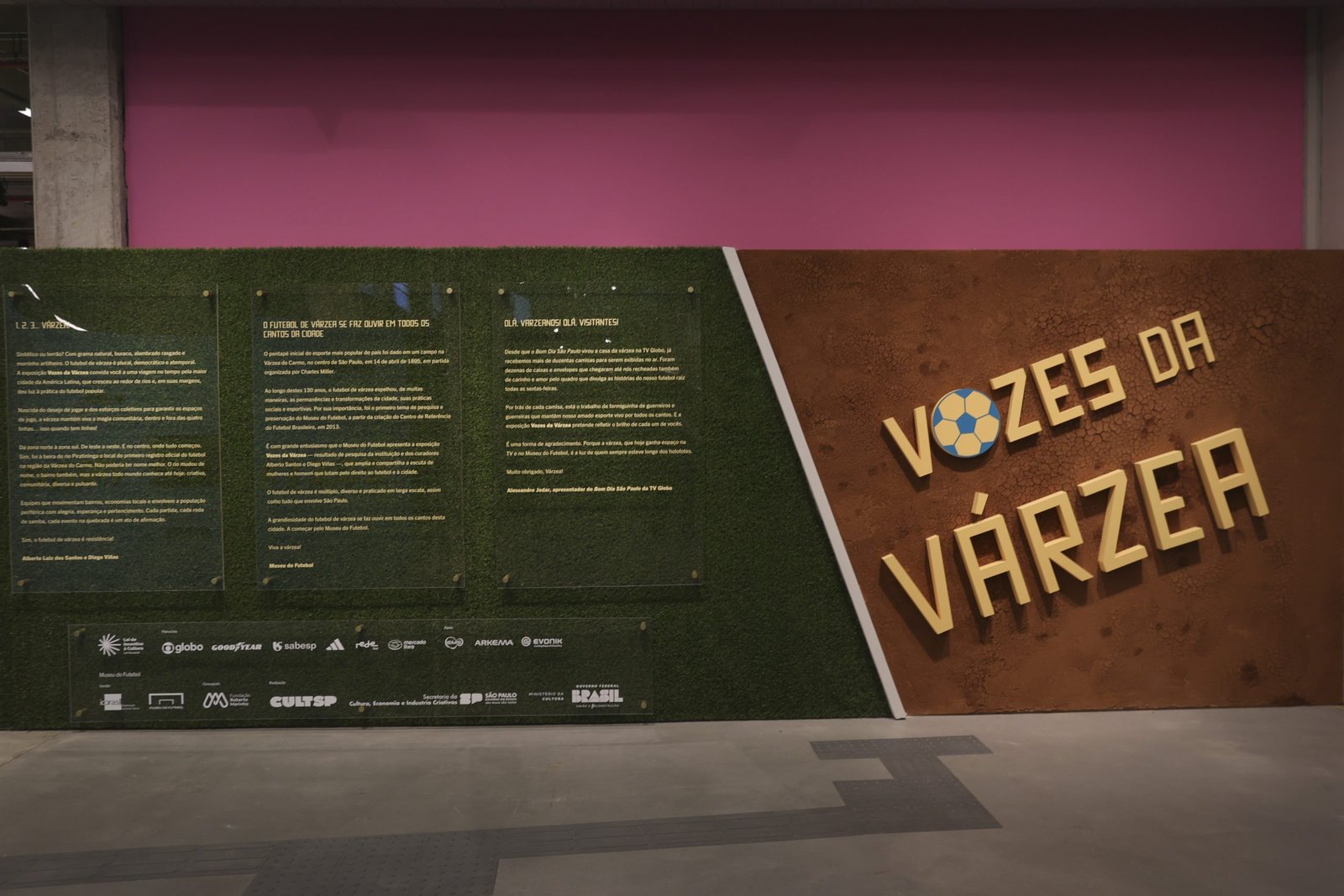 MF_Expo_Vozes_20241128_080 Painel de abertura da exposição mostrando a marca Vozes da Várzea sobre um fundo de terra seca e, ao lado, os textos sobre um fundo de grama sintética.