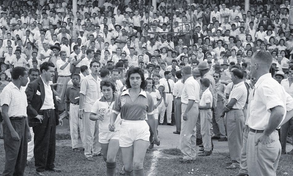 Imagem em preto e branco de mulheres vestidas com uniforme de futebol e torcedores ao fundo