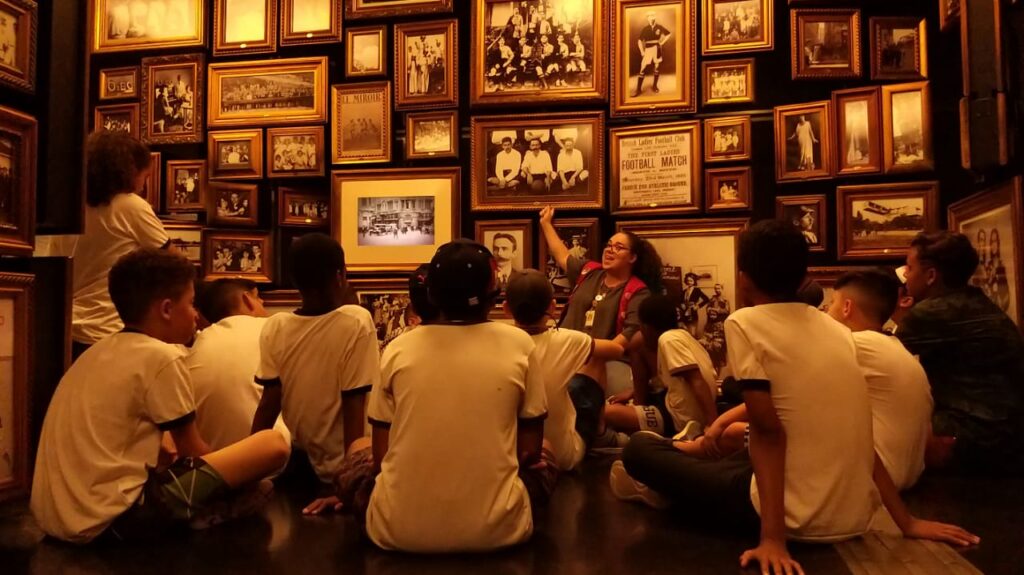 Crianças sentadas numa sala cercada por várias fotos antigas com molduras de madeira. Uma educadora com colete vermelho aponta uma das fotos e explica algo para o grupo.
