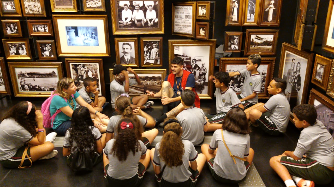 Grupo de crianças sentada no chão de uma sala cheia de fotos em preto e branco. Elas escutam um rapaz de colete vermelho, educador do museu, que também está sentado no chão.