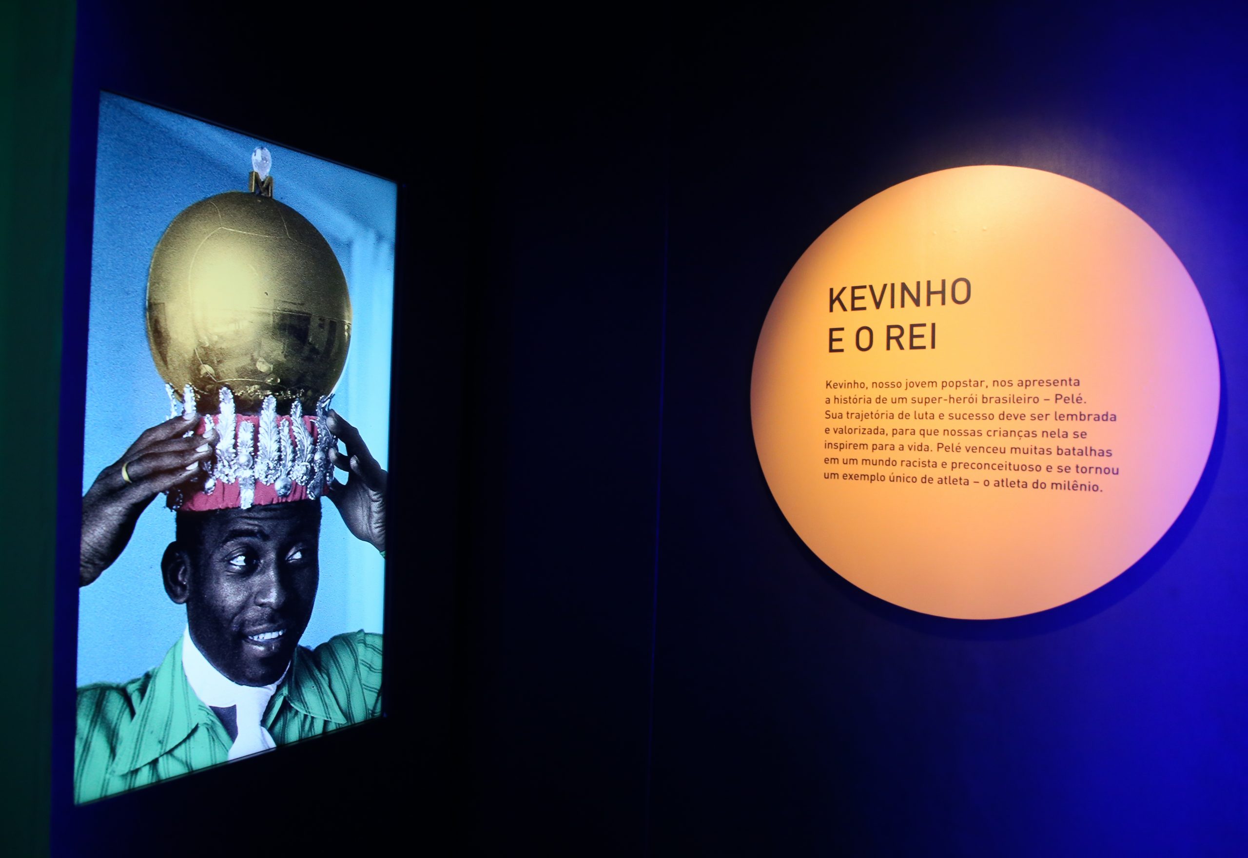 Em uma placa redonda está escrito "Kevinho e o Rei". Na tela de TV ao lado, aparece a imagem de Pelé colocando uma coroa, que tem uma bola em seu topo.