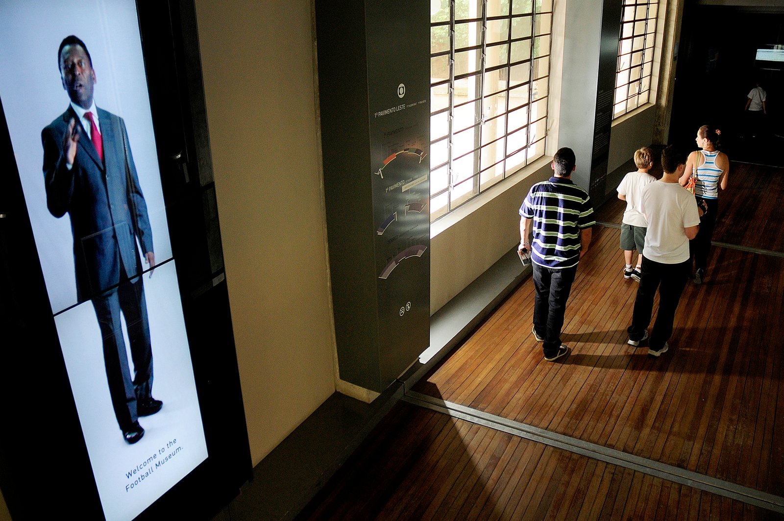 Parte de cima da entrada do Museu do Futebol. Recepção do Pelé em uma tela digital dando boas vindas aos visitantes. Depois dele, pessoas caminhando em direção ao Museu.