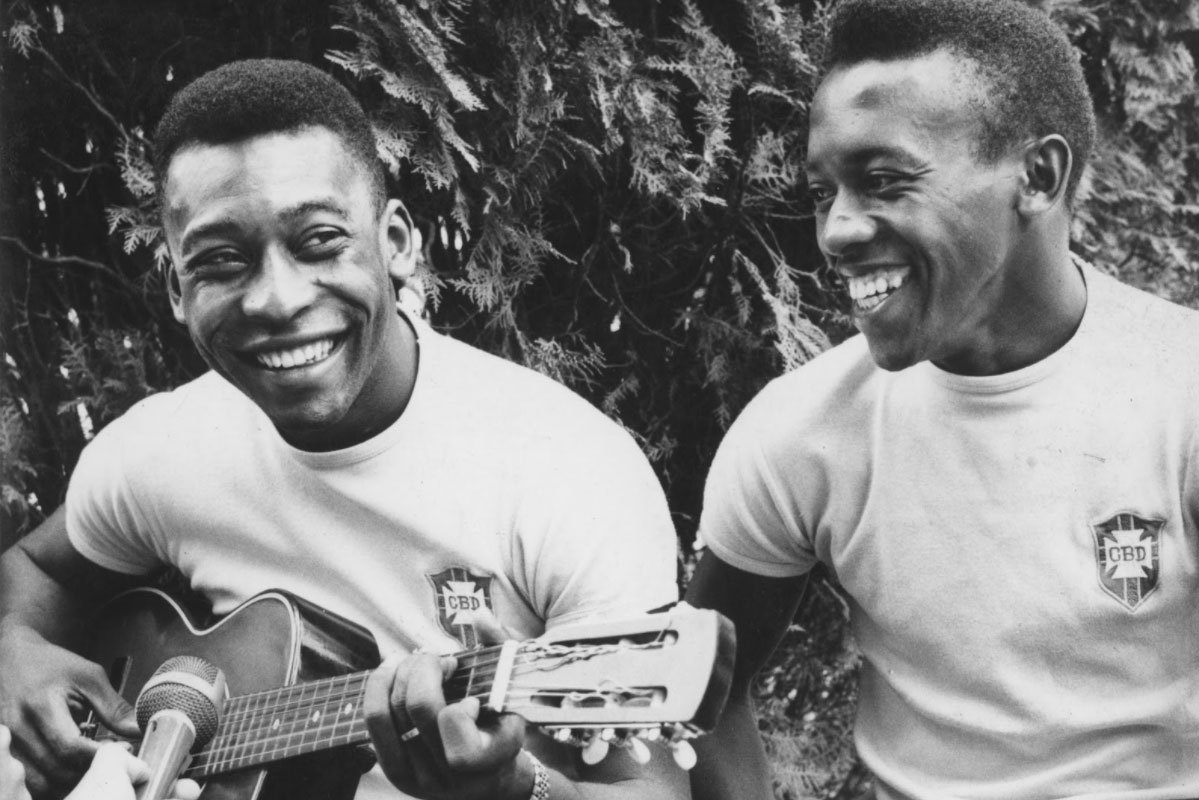 Pele toca violão ao lado de Ivair. Eles estão bastante sorridenters e vestem camisas da CBD.