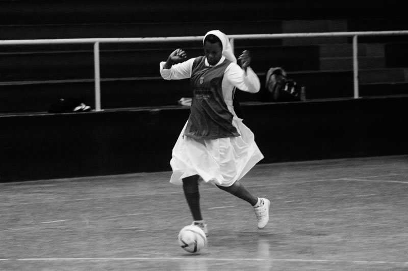 Uma mulher negra com vestes de freira domina uma bola com os pés em uma quadra. Sobre o hábito ela veste o colete do time.