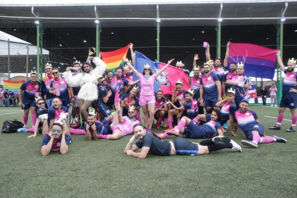Um time de futebol composto por homens e mulheres posa para foto em um gramado. Muitos dos rapazes estão deitados na grama e fazem poses engraçadas. Atrás deles, aparecem bandeiras do arco-íris, símbolo do orgulho LGBT.
