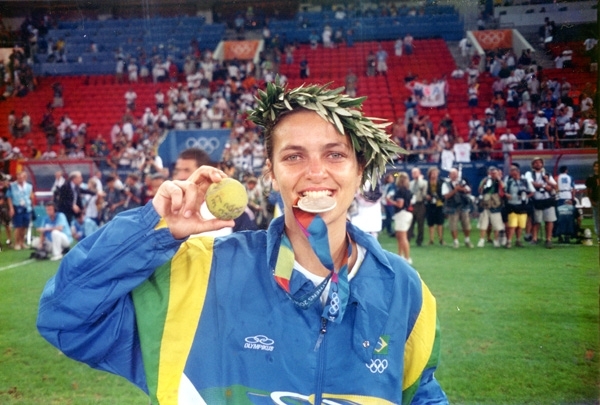 Foto de uma jogadora de futebol com agasalho olímpico mordendo uma medalha e com um ramo de folhas na cabeça, como uma coroa.