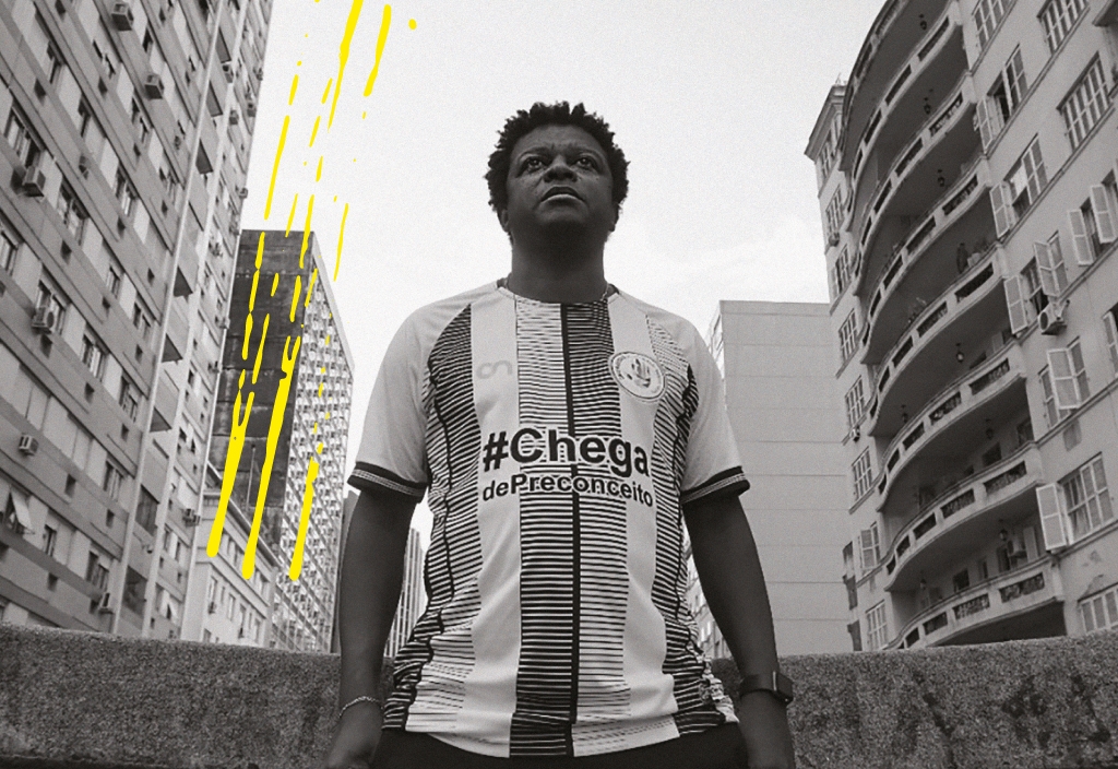 Marcelo Carvalho, um homem negro, numa foto em preto e branco em ambiente urbano, veste uma camisa onde está escrito "Chega de discriminação"