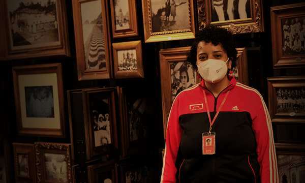 Educadora com agasalho do museu do futebol, de máscara, numa sala com vários quadros em preto e branco e molduras douradas.