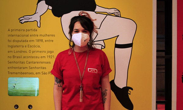 Educadora com camiseta vermelha do museu do futebol, com máscara, posa para foto na frente de uma placa amarela onde se vê desenhadas as pernas de uma jogadora de futebol