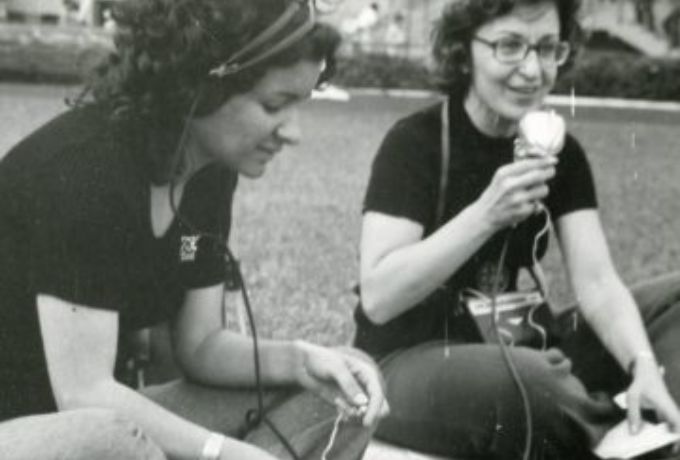 Foto em preto e branco de duas mulheres sentadas em um gramado, sendo que uma delas fala ao microfone de um gravador.