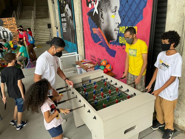 Crianças e adolescentes brincam numa mesa de pebolim na área externa do Museu do Futebol, enquanto várias pessoas circulam pelo espaço.