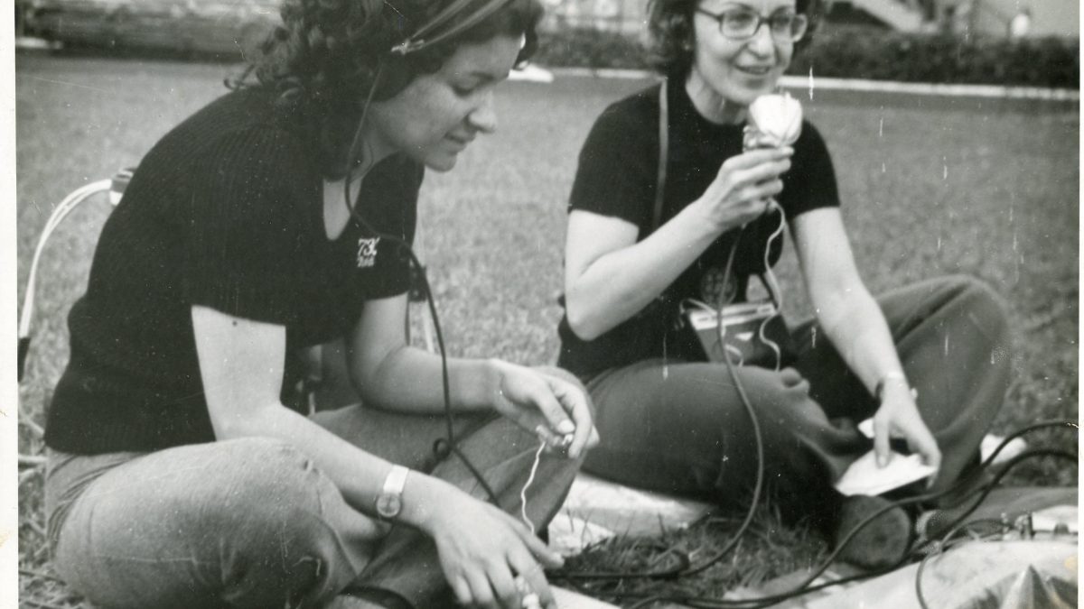 Foto em preto e branco de duas mulheres sentadas em um gramado, sendo que uma delas fala ao microfone de um gravador.