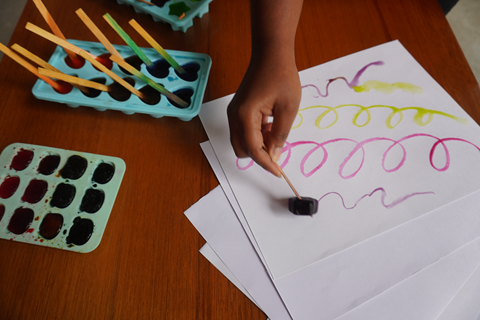 Uma mão aparece desenhando em papel com um dos gelos coloridos, segurando pelo palito. 