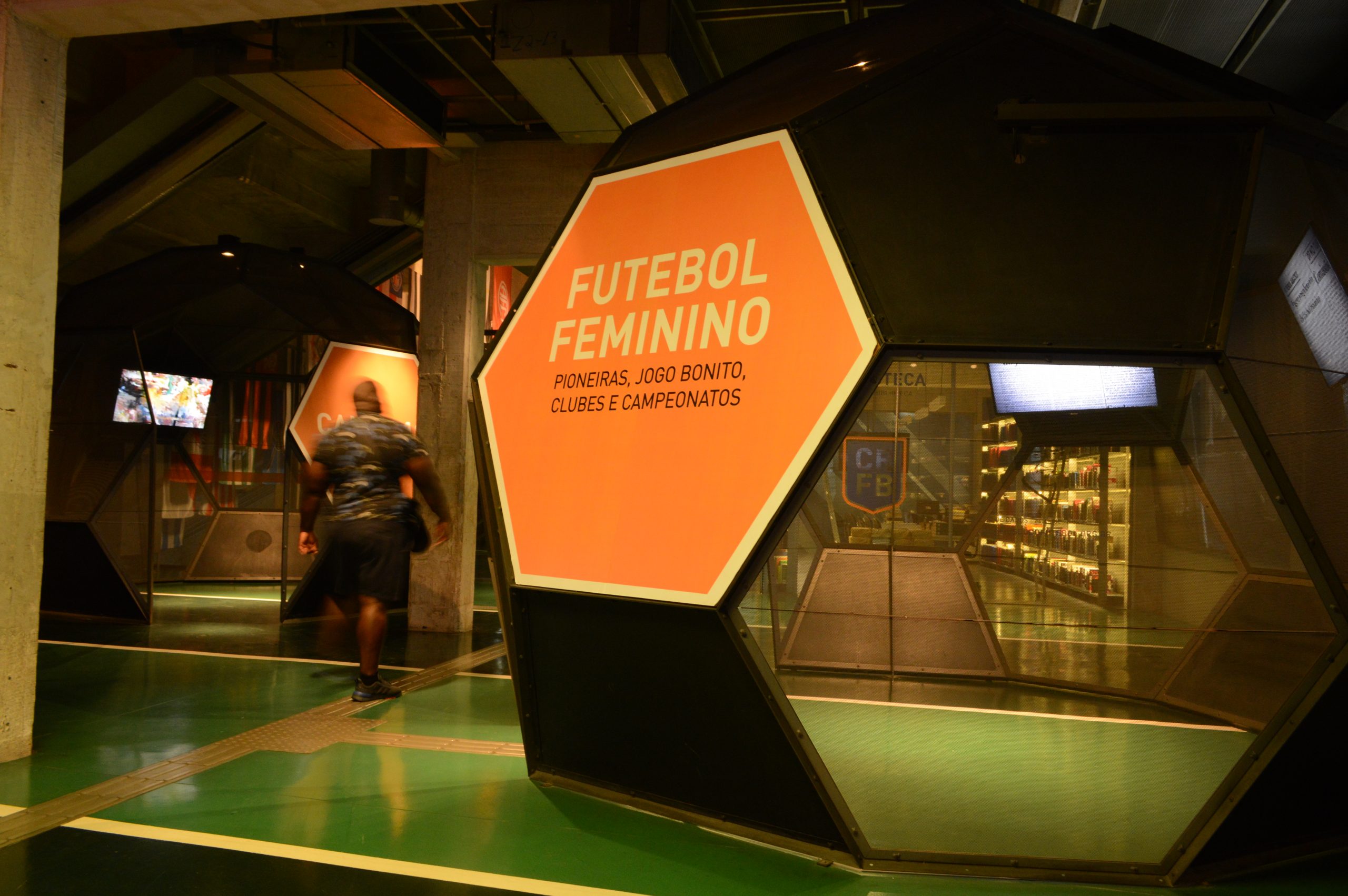 Foto de um ambiente do Museu do Futebol com uma estrutura metálica de formas hexagonais, dentro das quais há telas de TV. Na primeira delas lemos numa grande placa a frase "Futebol Feminino"